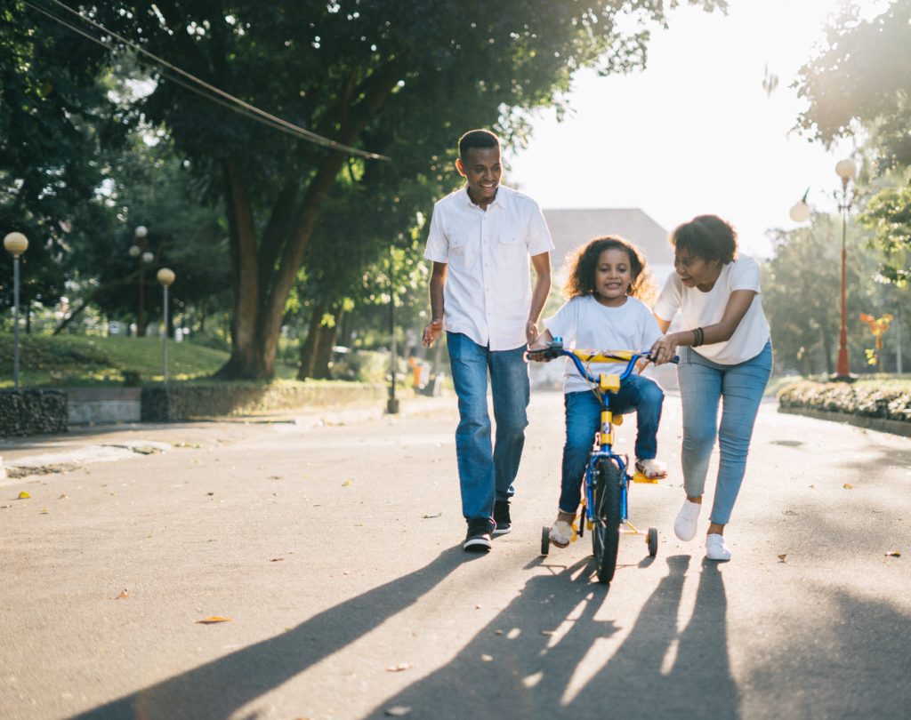 Two parents walk down the street next to their child who is learning to ride a bicycle