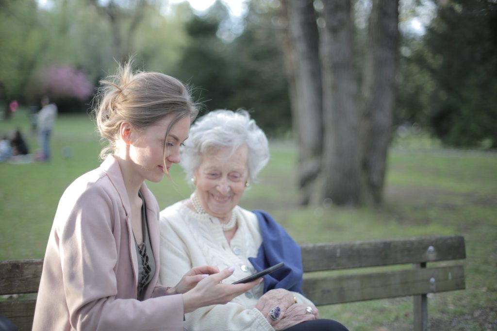 Two individuals sitting on a park bench side by side; on the right is a white-haired senior, while on the left is a younger person holding a smartphone.