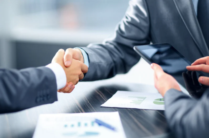 Two men in suits shaking hands across a table while another man is using a tablet